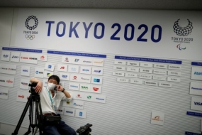 Seorang kamerawan mengenakan masker pelindung wajah menyusul wabah penyakit virus corona (COVID-19) di depan spanduk yang memperlihatkan logo-logo perusahaan dan sponsor Olimpiade dan Paralimpiade Tokyo 2020 di ruang media di Tokyo, Jepang, 17 Maret 2020. (REUTERS/ISSEI KATO)